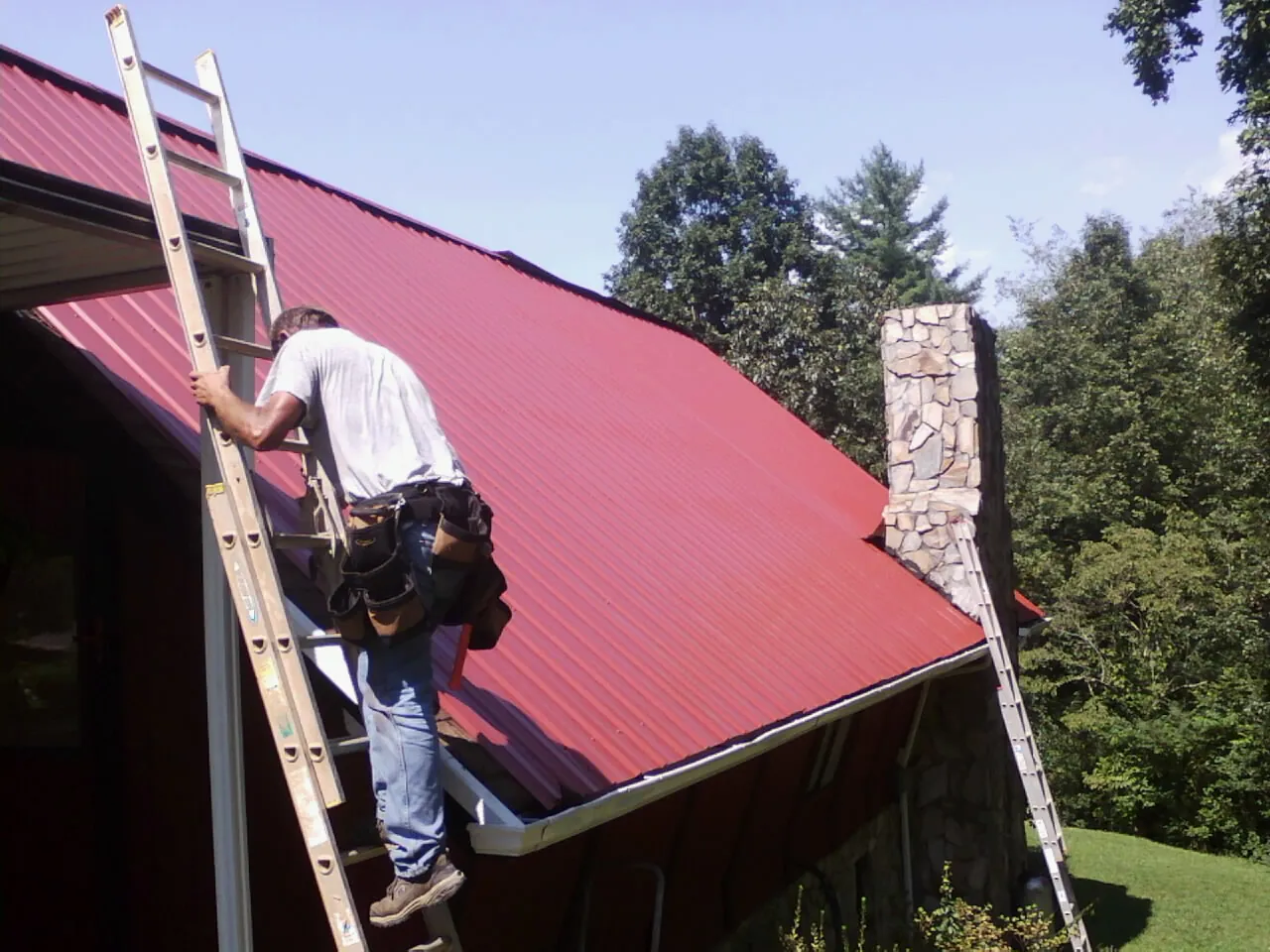 Roofer on a ladder beside a red metal roof and stone chimney — Soffit & Fascia Repair by Roofing Brothers in Shannon City